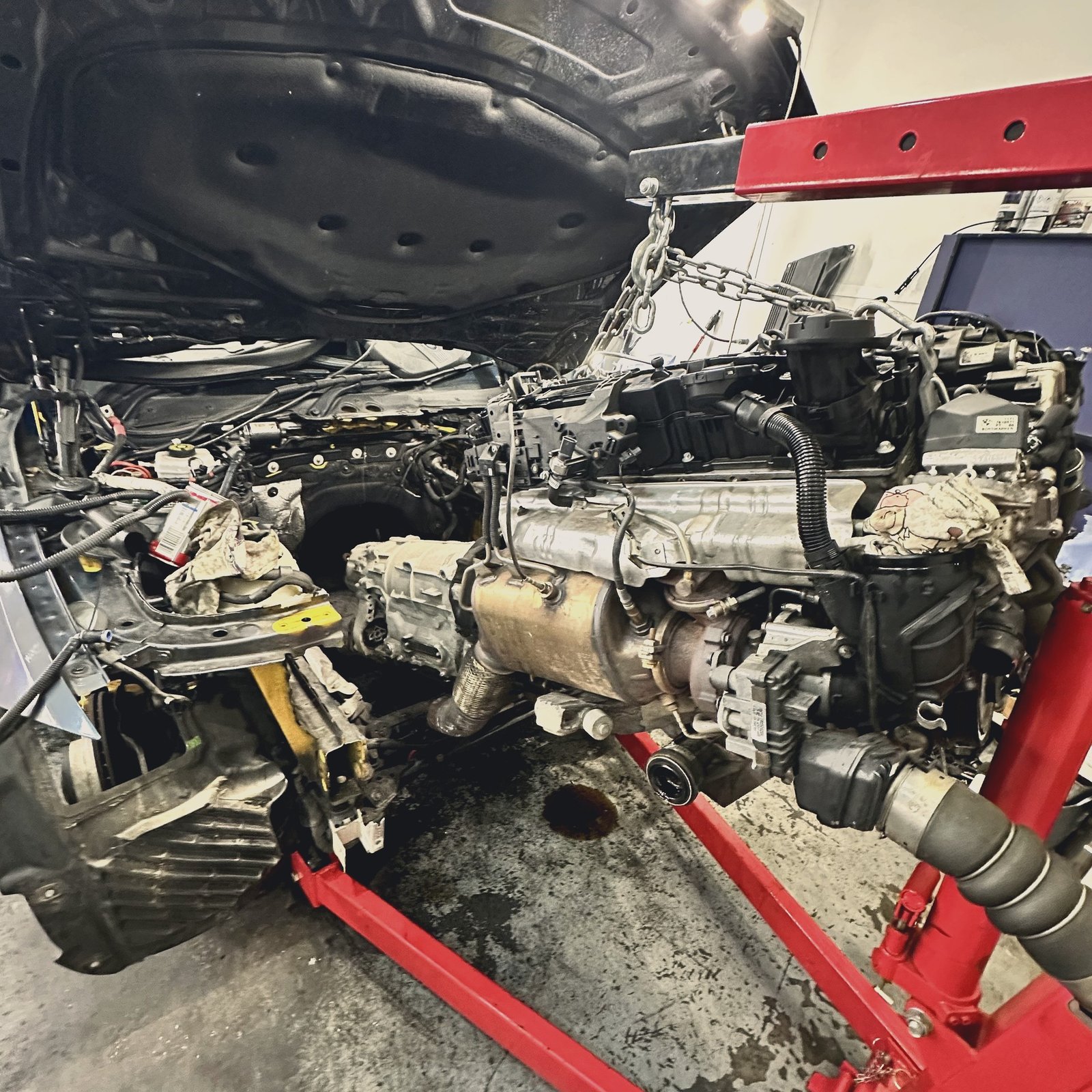 Engine and gearbox partially removed from a car, hanging from a workshop crane